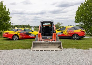 An eye-level shot features an orange Kubota skid steer facing away from the camera, positioned between two fleet vehicles on a grassy area. To the left is a yellow and red sedan, and to the right is a matching SUV, both prominently displaying "INC" branding and the text "CONCRETE & EXCAVATION SOLUTIONS" alongside a phone number. The machinery and vehicles are parked on a strip of green grass bordering a gravel lot. In the background, a small pond, lush green trees, and a white water tower are visible under a cloudy sky.