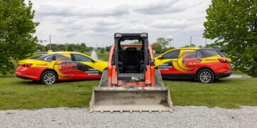 An eye-level shot features an orange Kubota skid steer facing away from the camera, positioned between two fleet vehicles on a grassy area. To the left is a yellow and red sedan, and to the right is a matching SUV, both prominently displaying "INC" branding and the text "CONCRETE & EXCAVATION SOLUTIONS" alongside a phone number. The machinery and vehicles are parked on a strip of green grass bordering a gravel lot. In the background, a small pond, lush green trees, and a white water tower are visible under a cloudy sky.