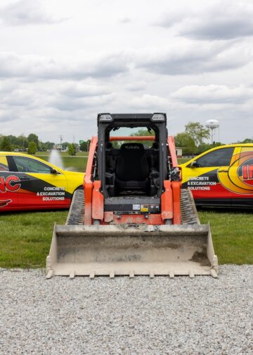 An eye-level shot features an orange Kubota skid steer facing away from the camera, positioned between two fleet vehicles on a grassy area. To the left is a yellow and red sedan, and to the right is a matching SUV, both prominently displaying "INC" branding and the text "CONCRETE & EXCAVATION SOLUTIONS" alongside a phone number. The machinery and vehicles are parked on a strip of green grass bordering a gravel lot. In the background, a small pond, lush green trees, and a white water tower are visible under a cloudy sky.