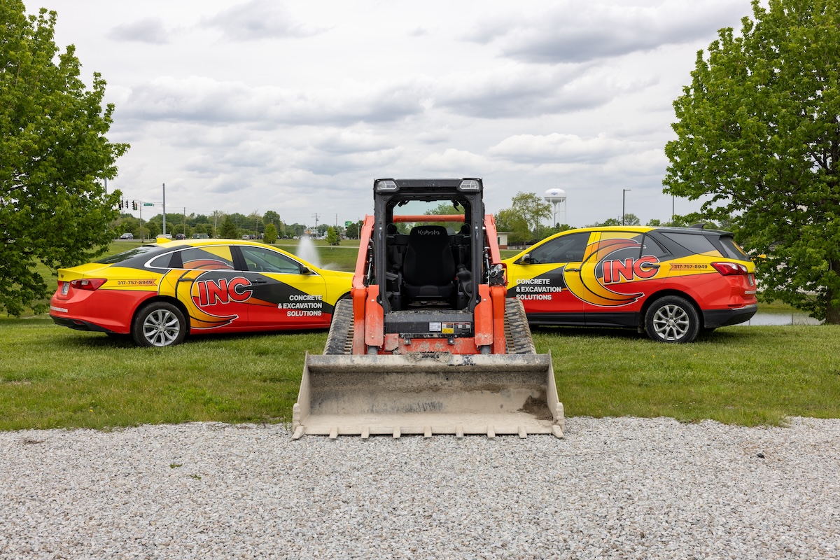 An eye-level shot features an orange Kubota skid steer facing away from the camera, positioned between two fleet vehicles on a grassy area. To the left is a yellow and red sedan, and to the right is a matching SUV, both prominently displaying "INC" branding and the text "CONCRETE & EXCAVATION SOLUTIONS" alongside a phone number. The machinery and vehicles are parked on a strip of green grass bordering a gravel lot. In the background, a small pond, lush green trees, and a white water tower are visible under a cloudy sky.