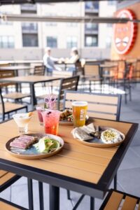 A bright outdoor patio at Sun King Brewery with several tables and chairs. In the foreground, a wooden table is spread with a feast: seared tuna, pita with dip, a pint of beer in a Sun King glass, and three colorful cocktails. In the blurred background, other diners enjoy the sun on the patio.