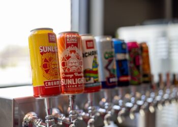A row of beer tap handles in a brewery, each topped with a colorful Sun King Brewery can. The first can is a yellow and red Sunlight Cream Ale. The second is an orange Orange Vanilla Sunlight Cream Ale. Other cans in the blurred background include Pachanga and Osito. The taps are mounted on a long, stainless steel bar.