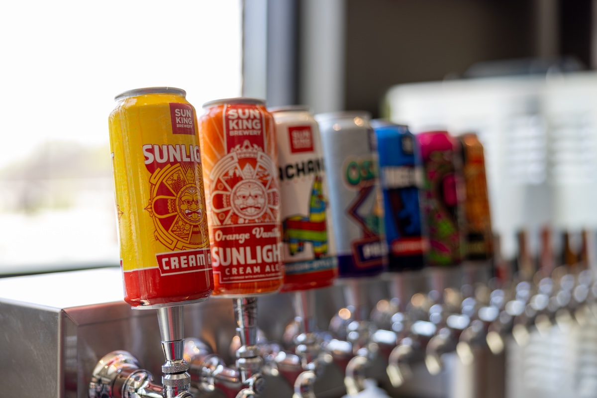 A row of beer tap handles in a brewery, each topped with a colorful Sun King Brewery can. The first can is a yellow and red Sunlight Cream Ale. The second is an orange Orange Vanilla Sunlight Cream Ale. Other cans in the blurred background include Pachanga and Osito. The taps are mounted on a long, stainless steel bar.