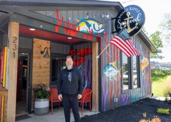 A man with a blonde beard and black jacket stands outside Broad Ripple Seafood in Indianapolis. The building is grey corrugated metal with colorful murals of tuna and coral. An American flag hangs over the entrance, and a circular "Broad Ripple Seafood Fish & Bar" sign is mounted on the wall. Two red chairs and a potted plant sit by the wooden entryway.