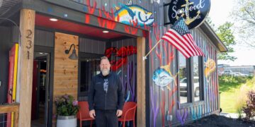 A man with a blonde beard and black jacket stands outside Broad Ripple Seafood in Indianapolis. The building is grey corrugated metal with colorful murals of tuna and coral. An American flag hangs over the entrance, and a circular "Broad Ripple Seafood Fish & Bar" sign is mounted on the wall. Two red chairs and a potted plant sit by the wooden entryway.