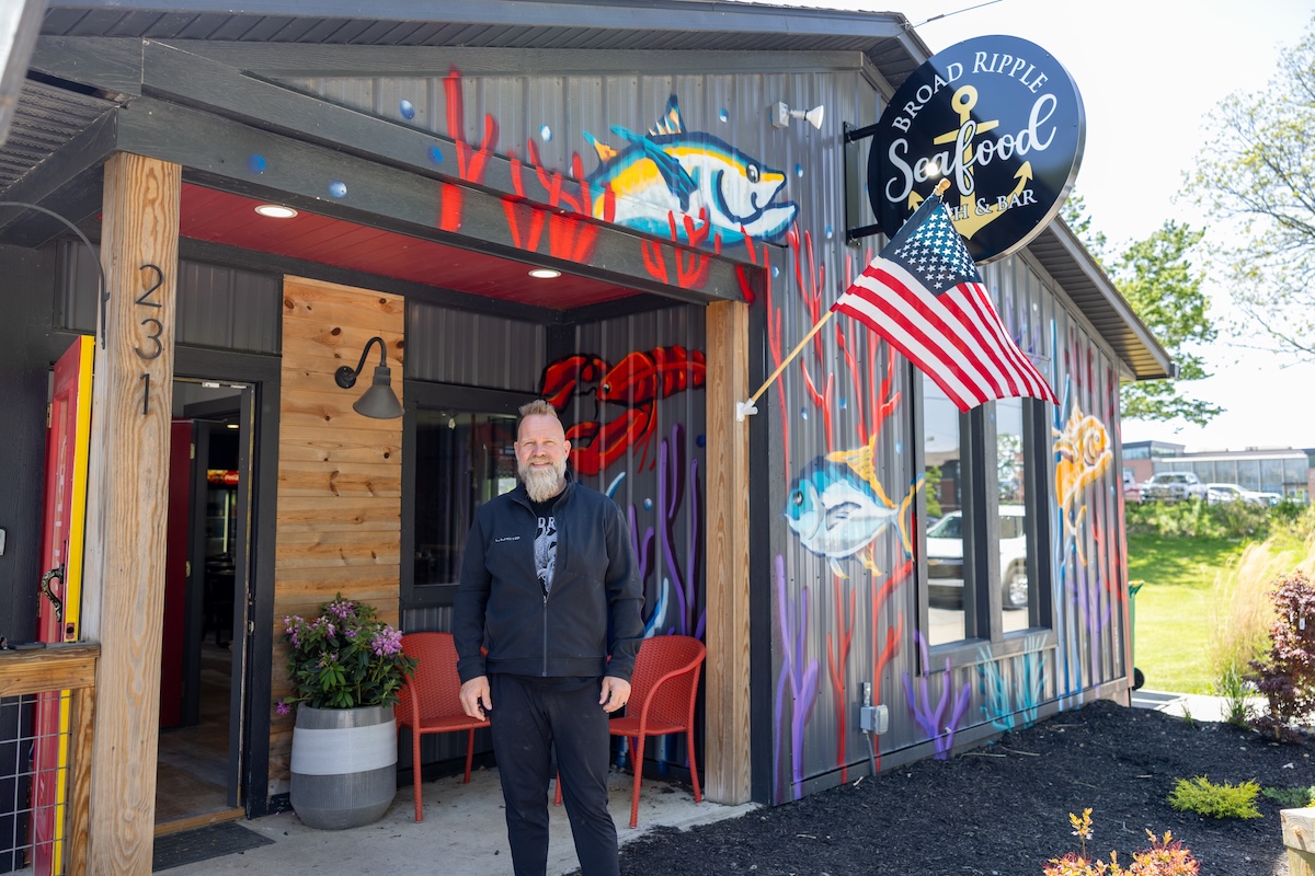 A man with a blonde beard and black jacket stands outside Broad Ripple Seafood in Indianapolis. The building is grey corrugated metal with colorful murals of tuna and coral. An American flag hangs over the entrance, and a circular "Broad Ripple Seafood Fish & Bar" sign is mounted on the wall. Two red chairs and a potted plant sit by the wooden entryway.
