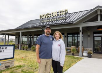 A man and woman stand smiling in front of a modern grey building with a black metal roof. A sign on the roof reads "PAWS ON TAP: TAPROOM & BEER GARDEN." To the left, a marquee sign says "OPENING MAY." The building features large glass windows and an outdoor patio area with wooden tables.