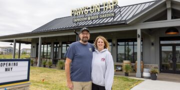 A man and woman stand smiling in front of a modern grey building with a black metal roof. A sign on the roof reads "PAWS ON TAP: TAPROOM & BEER GARDEN." To the left, a marquee sign says "OPENING MAY." The building features large glass windows and an outdoor patio area with wooden tables.