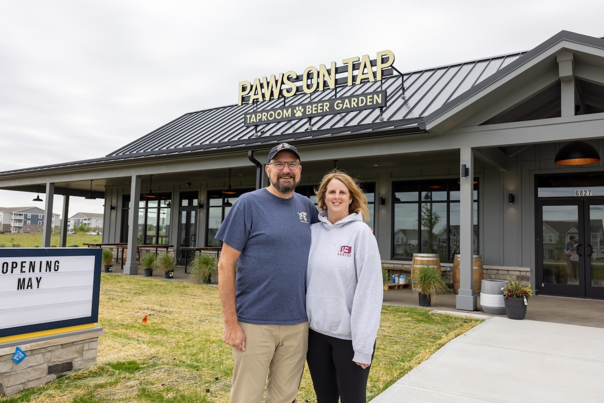 A man and woman stand smiling in front of a modern grey building with a black metal roof. A sign on the roof reads "PAWS ON TAP: TAPROOM & BEER GARDEN." To the left, a marquee sign says "OPENING MAY." The building features large glass windows and an outdoor patio area with wooden tables.
