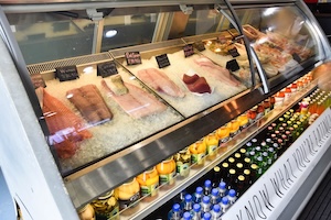 A bright, glass-fronted refrigerated display case at Broad Ripple Seafood Market. The top section features several varieties of fresh fish fillets, including salmon and tuna, resting on a bed of crushed ice with small black price tags. Below the fish, rows of bottled juices, sparkling waters, and sodas are neatly organized. The floor-level trim of the case features the shop's motto: "KNOW WHAT YOU'RE EATING."