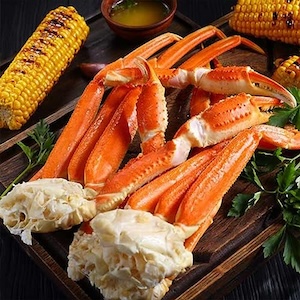 A close-up of steamed snow crab legs served on a dark wooden board. The crab legs are bright orange and white, accompanied by ears of grilled corn on the cob and fresh parsley sprigs. A small bowl of melted butter for dipping sits in the background.