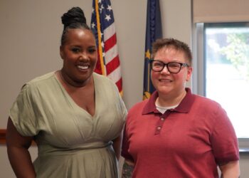 A medium-shot portrait of two women smiling side-by-side in an office or government building. The woman on the left has her hair in a high braided bun and wears a light green v-neck dress. The woman on the right wears black-rimmed glasses and a red polo shirt. Behind them, an American flag and an Indiana state flag stand on poles. A bright window is visible in the background to the right.