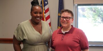A medium-shot portrait of two women smiling side-by-side in an office or government building. The woman on the left has her hair in a high braided bun and wears a light green v-neck dress. The woman on the right wears black-rimmed glasses and a red polo shirt. Behind them, an American flag and an Indiana state flag stand on poles. A bright window is visible in the background to the right.