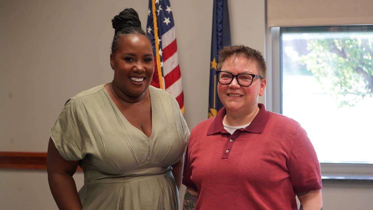 A medium-shot portrait of two women smiling side-by-side in an office or government building. The woman on the left has her hair in a high braided bun and wears a light green v-neck dress. The woman on the right wears black-rimmed glasses and a red polo shirt. Behind them, an American flag and an Indiana state flag stand on poles. A bright window is visible in the background to the right.