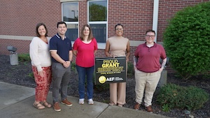 A group of five smiling professionals stands outside a brick building, posing for a commemorative photo. The person in the center holds a black sign with bold yellow and white text that reads, "PROUD GRANT RECIPIENT," featuring the "AEF" (Anderson Education Foundation) logo at the bottom. The group is dressed in business-casual attire, including sweaters, polo shirts, and slacks, and they are positioned on a sidewalk next to a manicured green bush.