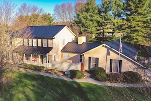 An aerial, wide-angle shot of a large two-story residential home with a tan exterior and a newly installed black metal roof. The property features a long covered front porch on the main two-story structure, which connects via a central walkway to a single-story wing with black decorative shutters. A manicured green lawn with low landscaping bushes slopes down in the foreground, while tall green pine trees and bare seasonal trees frame the property against a clear blue sky.