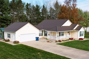 A high-angle exterior shot of a single-story ranch-style house with white siding and a new dark brown metal roof. The home features an attached two-car garage with a wide concrete driveway in the foreground. A covered front porch with white railings wraps around the entryway, bordered by neatly landscaped garden beds with small shrubs and red mulch. The property is surrounded by a green grass lawn, with dense green and autumn-colored trees in the background under a cloudy sky.