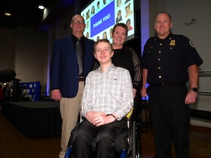 A young man in a wheelchair poses indoors alongside three adults, including a man in a blue blazer, a woman, and a police officer or first responder in a dark uniform. A presentation screen displaying a "Thank You" message and a grid of faces is illuminated in the background.