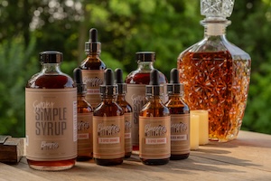 An assortment of brown glass bottles with labels from Bluegrass Bitters sits on a wooden outdoor surface. A large, texturized glass decanter filled with an amber liquid and small decorative elements are arranged behind the bottles under soft, natural lighting.