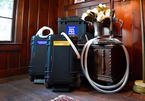 An indoor, low-angle shot of three portable MagnaWave PEMF equine therapy machines housed in rugged black cases, arranged together on a rustic wooden floor. The center machine features a large blue label that reads "MAGNAWAVE SOL PRO". The machines stand against a wood-paneled corner next to an antique wooden side table. The table features intricate carved pillars and a glass vase filled with white artificial roses on its top surface.