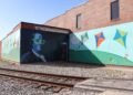A large, two-panel mural on the side of a red brick building next to railroad tracks. One panel features a stylized portrait of Benjamin Franklin wearing bright neon green glasses. The adjoining panel shows a whimsical scene of colorful kites flying over rolling green hills under a blue sky with white clouds.