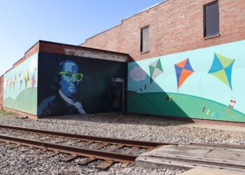 A large, two-panel mural on the side of a red brick building next to railroad tracks. One panel features a stylized portrait of Benjamin Franklin wearing bright neon green glasses. The adjoining panel shows a whimsical scene of colorful kites flying over rolling green hills under a blue sky with white clouds.