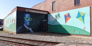 A large, two-panel mural on the side of a red brick building next to railroad tracks. One panel features a stylized portrait of Benjamin Franklin wearing bright neon green glasses. The adjoining panel shows a whimsical scene of colorful kites flying over rolling green hills under a blue sky with white clouds.