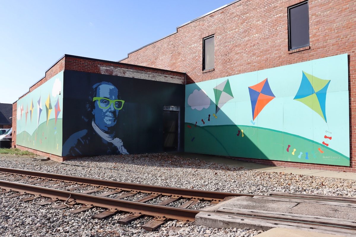 A large, two-panel mural on the side of a red brick building next to railroad tracks. One panel features a stylized portrait of Benjamin Franklin wearing bright neon green glasses. The adjoining panel shows a whimsical scene of colorful kites flying over rolling green hills under a blue sky with white clouds.