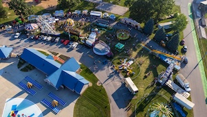 An aerial drone shot of a small carnival or county fair during the day. The scene features various amusement rides, including a small roller coaster, a spinning ride, and several game booths. Colorful tents and trailers are scattered throughout the grassy and paved areas. A large blue-roofed building and a parking lot are visible on the left side of the frame.