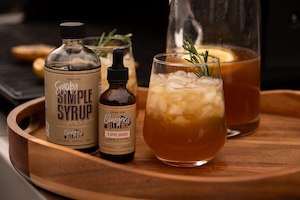 A round wooden serving tray holds an iced cocktail garnished with a fresh rosemary sprig. Next to the glass sit two brown bottles: a larger bottle labeled "Simple Syrup" and a smaller dropper bottle of Bluegrass Bitters, with a filled pitcher visible in the background.