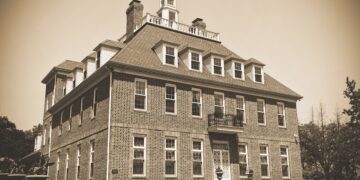 A sepia-toned photograph of the Coxhall Mansion, a large, three-story brick estate with Colonial Revival architecture. The building features a prominent cupola with a weather vane on top, numerous dormer windows on the roof, and a small iron balcony above the main entrance. Manicured landscaping and low shrubs line the front of the building.