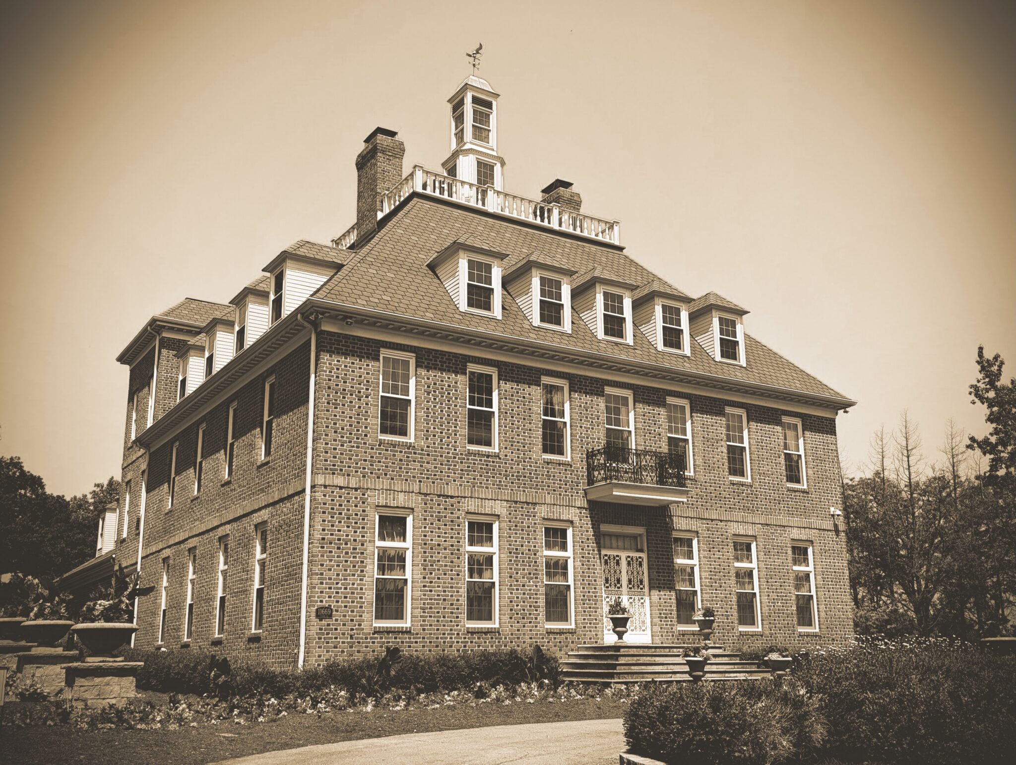 A sepia-toned photograph of the Coxhall Mansion, a large, three-story brick estate with Colonial Revival architecture. The building features a prominent cupola with a weather vane on top, numerous dormer windows on the roof, and a small iron balcony above the main entrance. Manicured landscaping and low shrubs line the front of the building.