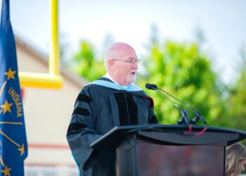 An older, bald man with glasses wearing a black doctoral graduation gown with velvet stripes speaks at a dark wooden podium during an outdoor ceremony. The Indiana state flag and a yellow football goalpost stand in the blurred background.