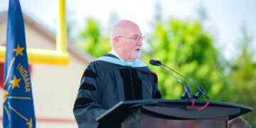 An older, bald man with glasses wearing a black doctoral graduation gown with velvet stripes speaks at a dark wooden podium during an outdoor ceremony. The Indiana state flag and a yellow football goalpost stand in the blurred background.