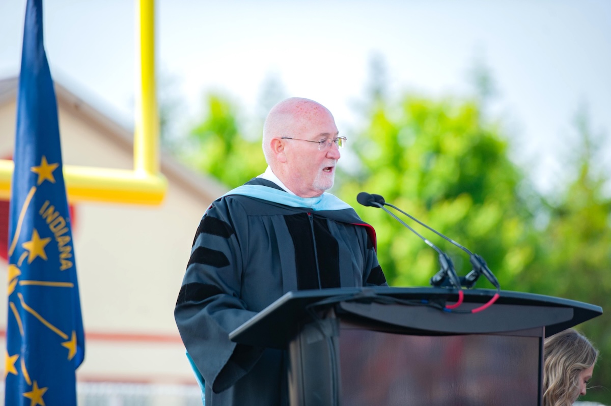 An older, bald man with glasses wearing a black doctoral graduation gown with velvet stripes speaks at a dark wooden podium during an outdoor ceremony. The Indiana state flag and a yellow football goalpost stand in the blurred background.