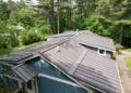 A high-angle, downward view of a multi-gabled residential roof finished with dark brown standing seam metal panels. The home features slate-blue horizontal siding with white trim accents and visible roof ventilation pipes. A stone walkway curves along the right side of the house, surrounded by dense green shrubs and a background of tall, mature pine and deciduous trees under soft daylight.