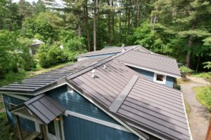 A high-angle, downward view of a multi-gabled residential roof finished with dark brown standing seam metal panels. The home features slate-blue horizontal siding with white trim accents and visible roof ventilation pipes. A stone walkway curves along the right side of the house, surrounded by dense green shrubs and a background of tall, mature pine and deciduous trees under soft daylight.