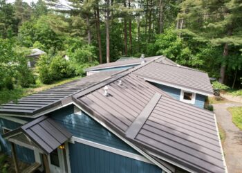 A high-angle, downward view of a multi-gabled residential roof finished with dark brown standing seam metal panels. The home features slate-blue horizontal siding with white trim accents and visible roof ventilation pipes. A stone walkway curves along the right side of the house, surrounded by dense green shrubs and a background of tall, mature pine and deciduous trees under soft daylight.