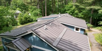 A high-angle, downward view of a multi-gabled residential roof finished with dark brown standing seam metal panels. The home features slate-blue horizontal siding with white trim accents and visible roof ventilation pipes. A stone walkway curves along the right side of the house, surrounded by dense green shrubs and a background of tall, mature pine and deciduous trees under soft daylight.