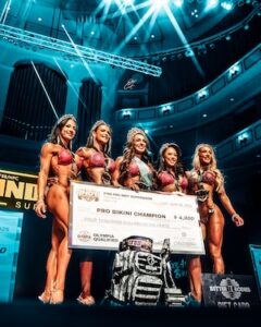 Five female fitness competitors in sparkling red bikinis stand together smiling on a stage beneath bright blue spotlights. The middle competitor wears a tiara and a winner's sash while holding a large prize check that reads "Pro Bikini Champion." A camouflage backpack and various trophies rest on the floor in front of them against the background of a large auditorium hall.