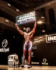 A male physique competitor stands on stage, smiling and raising a large sign overhead that reads "Overall Champion Optimize360." He wears patterned board shorts, showcasing a highly muscular and defined upper body. A camouflage backpack and a trophy sit on the stage floor beside him, with a dark background featuring "Wolfpak" and "Indy Pro" sponsor logos under bright overhead lighting.