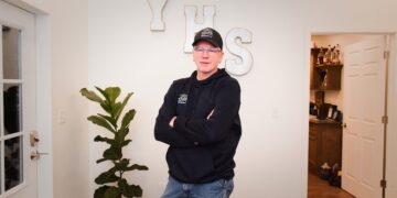A medium full-length indoor portrait shows a man standing in an office or reception area. He is wearing a black baseball cap, glasses, a black hoodie with a small blue and white logo on the left chest, and blue jeans. He has his arms crossed and is smiling slightly at the camera. Behind him, three large, decorative white block letters spelling "YHS" are mounted on a plain white wall. A potted fiddle-leaf fig plant stands to his left near a white entry door. To the right, an open doorway reveals a glimpse of an adjoining room with wooden cabinetry and shelves holding bottles.