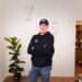 A medium full-length indoor portrait shows a man standing in an office or reception area. He is wearing a black baseball cap, glasses, a black hoodie with a small blue and white logo on the left chest, and blue jeans. He has his arms crossed and is smiling slightly at the camera. Behind him, three large, decorative white block letters spelling "YHS" are mounted on a plain white wall. A potted fiddle-leaf fig plant stands to his left near a white entry door. To the right, an open doorway reveals a glimpse of an adjoining room with wooden cabinetry and shelves holding bottles.