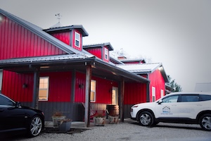 An exterior commercial shot shows a multi-gabled, barn-style building with vibrant red vertical metal siding and a dark gray lower foundation. The structure features three matching roof dormers, each topped with a small white cupola and weather vane, along a metal roof line. A covered walkway with rustic wooden support columns runs along the side entrance. In the foreground, a gravel driveway holds a dark car on the left and a white crossover SUV on the right, which features a house-shaped business logo on its front door. The background is a stark, hazy gray sky.
