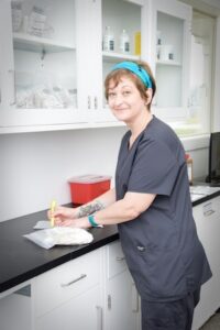 A woman with short hair and a bright blue headband stands behind a black laboratory counter, smiling directly at the camera. She wears dark gray medical scrubs and has a tattoo on her forearm. In her hand, she holds a yellow pen, using it to write on a clear, sealed package resting on the surface. The background features white cabinetry, including upper cabinets with glass doors filled with medical supplies and bottles, a small red sharps container, and a lower row of drawers.