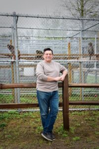 A full-length outdoor portrait of a person with short dark hair, wearing a long-sleeved beige crewneck sweater, blue jeans, and dark shoes. The person is smiling and leaning their arm on a dark brown wooden post-and-rail fence. In the background, three monkeys are visible sitting on wooden planks inside a large, high chain-link cage structure. The ground is a mix of sparse green grass and dirt under a hazy, white sky with bare tree tops visible at the top right.