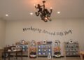 An interior shot of a brightly lit retail store display wall. The text "Monkeying Around Gift Shop" is painted in a dark, whimsical font in a gentle arch across the center of a plain beige wall. Below the sign, five wooden shelf units hold neatly stacked apparel, baseball caps, and plush monkey toys. Hanging from the ceiling in the upper-center is a unique dark metal chandelier featuring sculpted monkeys climbing on the arms between candle-style light bulbs. A round wooden table on the left displays photo frames, and a warm, clean floor runs beneath the displays.