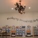 An interior shot of a brightly lit retail store display wall. The text "Monkeying Around Gift Shop" is painted in a dark, whimsical font in a gentle arch across the center of a plain beige wall. Below the sign, five wooden shelf units hold neatly stacked apparel, baseball caps, and plush monkey toys. Hanging from the ceiling in the upper-center is a unique dark metal chandelier featuring sculpted monkeys climbing on the arms between candle-style light bulbs. A round wooden table on the left displays photo frames, and a warm, clean floor runs beneath the displays.