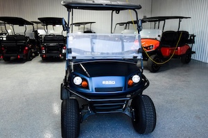 A dark metallic blue E-Z-GO golf cart faces forward in the center of a showroom. It features a clear windshield, round headlights with orange turn signals, a black roof, and rugged black tires. The E-Z-GO logo is visible on the front cowl. The vehicle sits on a gray speckled epoxy floor inside a garage or warehouse with white corrugated metal walls. Several other golf carts, including black and bright orange models, are parked in rows behind it.
