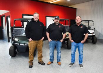 Three middle-aged men stand in a row facing the camera with their hands in their pockets inside a vehicle showroom. The man on the left wears glasses, a black hoodie, tan pants, and work boots. The center man wears a black baseball cap, a black polo shirt, blue jeans, and brown boots. The man on the right wears a black polo shirt, blue jeans, and gray slip-on shoes. Both polo shirts and the hoodie feature a small red and white "GT" logo on the left chest. Behind them, three modern golf carts—one dark green, one tan, and one white—are parked on a gray speckled epoxy floor against a vibrant red and dark gray wall.